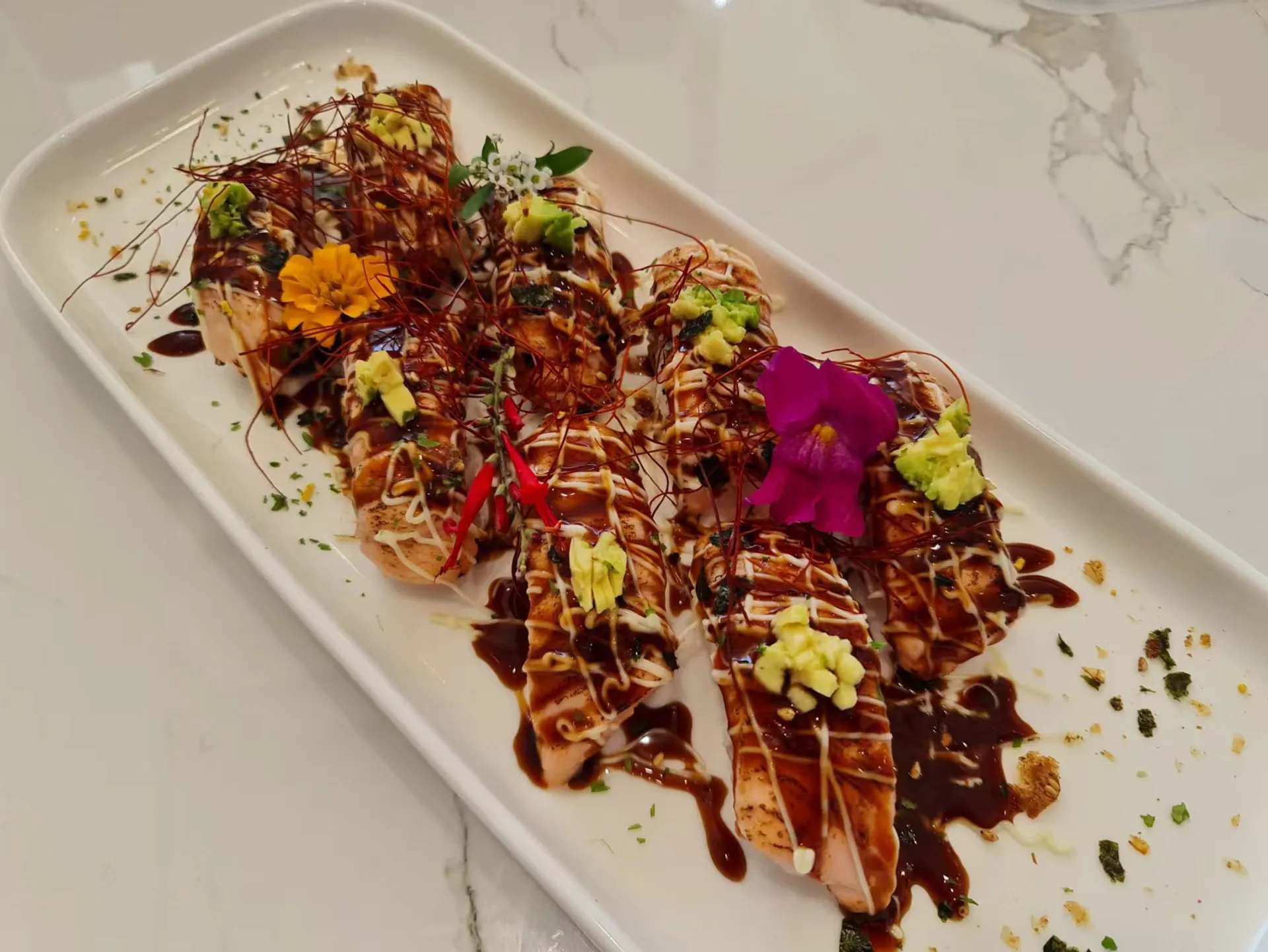 Aburi salmon rolls with edible flowers, avocado and chilli threads on white marble plate