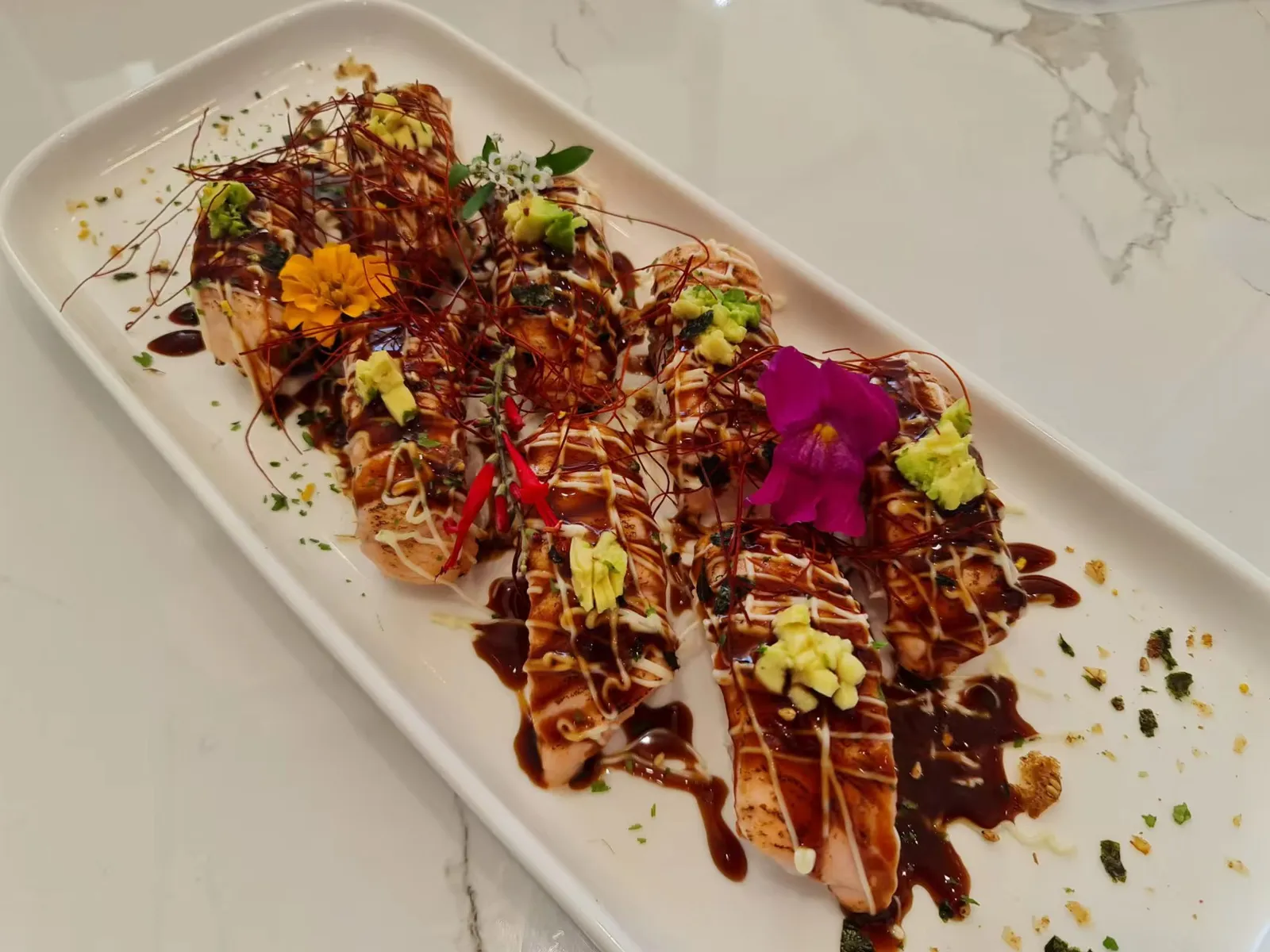 Aburi salmon rolls with edible flowers, avocado and chilli threads on white marble plate