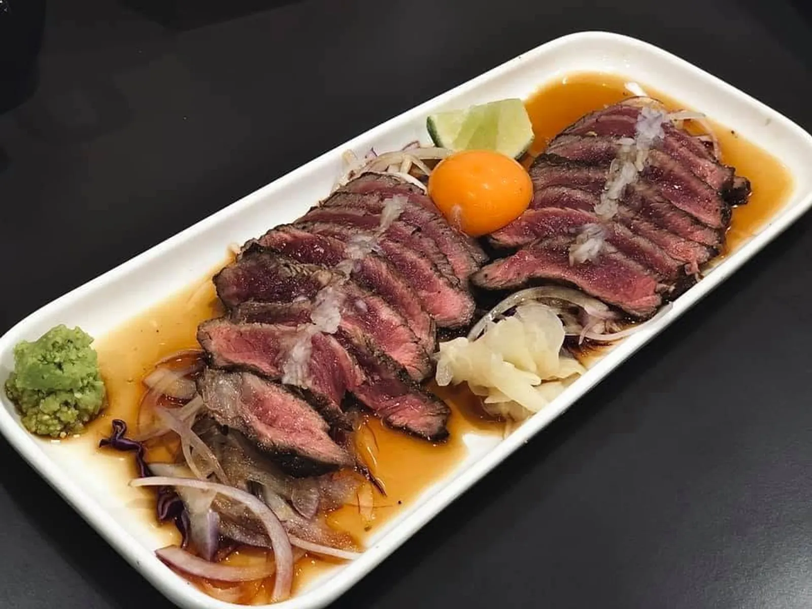 Sliced beef tataki on white plate with wasabi, pickled ginger and amber sauce, dark background