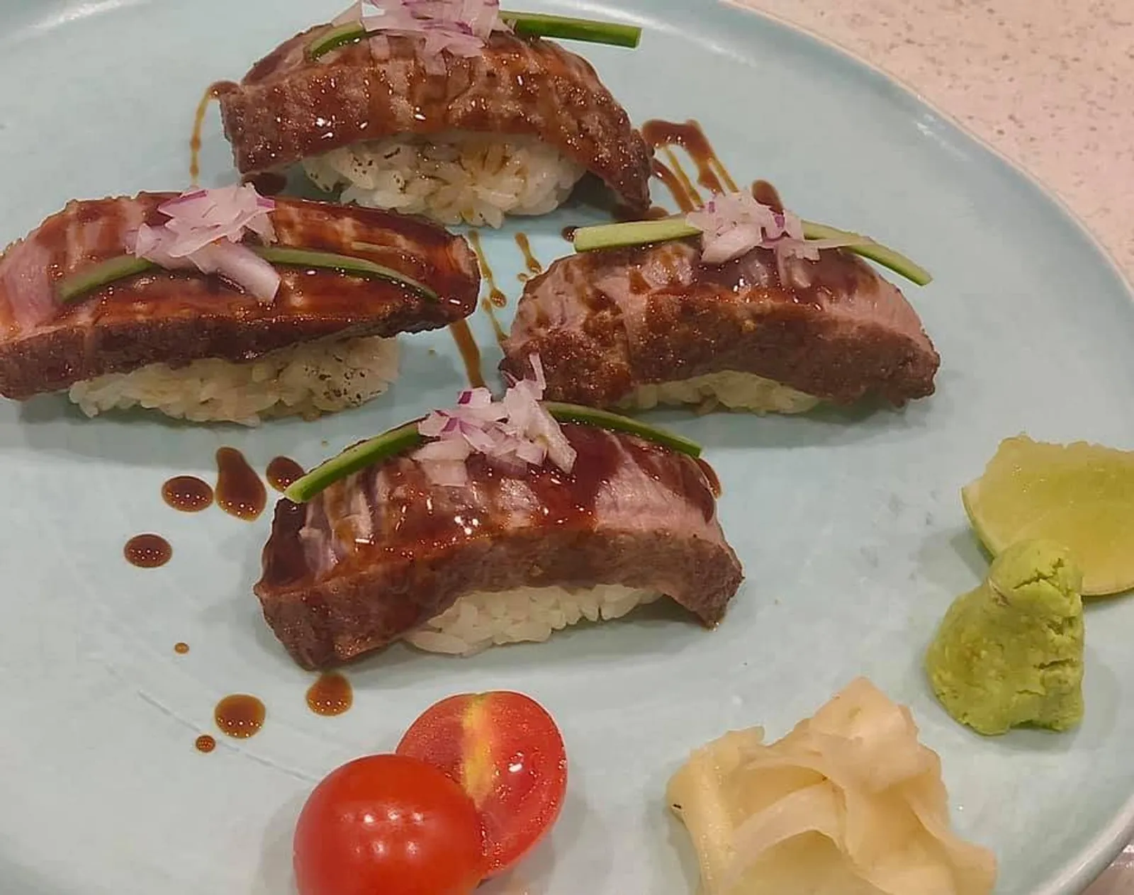 Four wagyu beef nigiri on blue-grey plate with teriyaki glaze, cucumber and pickled ginger